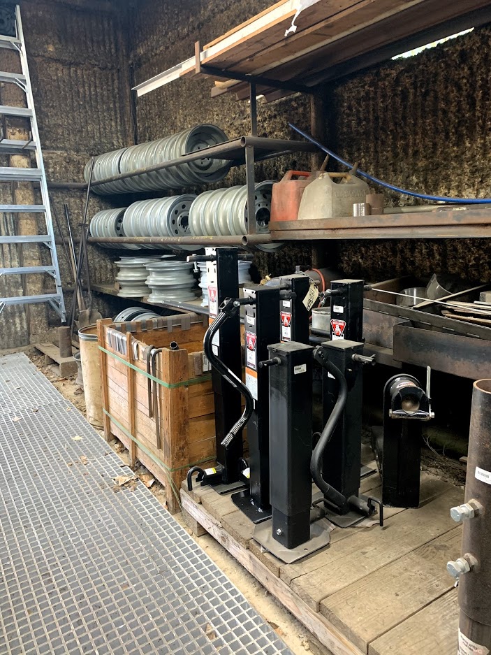 Heavy-duty black trailer tongue jacks on a pallet with trailer rims on an upper rack at Riggs Machine & Welding in Stephenville, TX