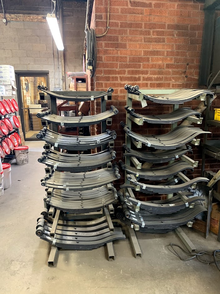 Two towers of trailer leaf spring assemblies stacked on stands in the Riggs Machine & Welding shop, Stephenville, TX