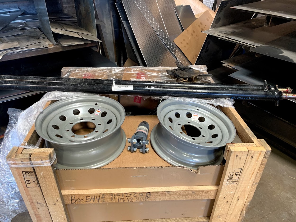 Trailer axle kit on a wooden crate at Riggs Machine & Welding in Stephenville, TX — two 8-lug silver rims, axle tubes, and a coupler with plate steel behind