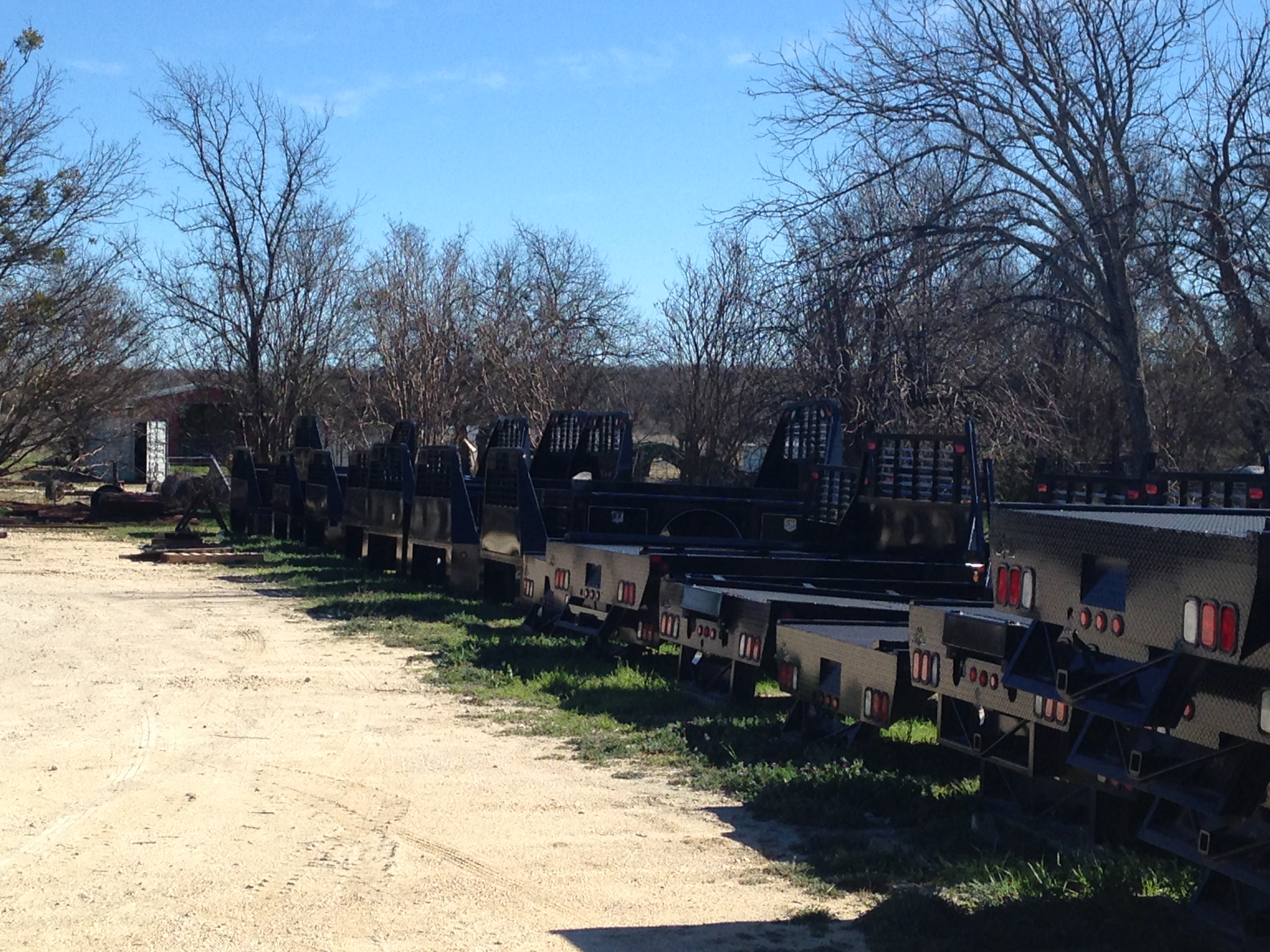 Row of black J-I MFG flatbed truck beds with headache racks lined up in the yard at Riggs Machine & Welding, Stephenville, TX